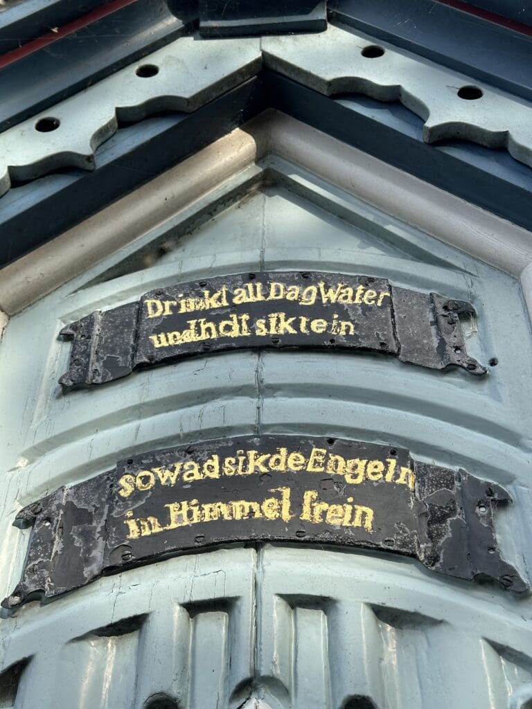 Plattdeutsche Verse von Klaus Groth zieren in goldenen Buchstaben das Brunnenhäuschen der Friedrichstädter Marktpumpe, Text auf der Südseite: "Drinkt all Drink all Dag Water / und holt sik rein / sowad sik de Engeln / in Himmel frein"