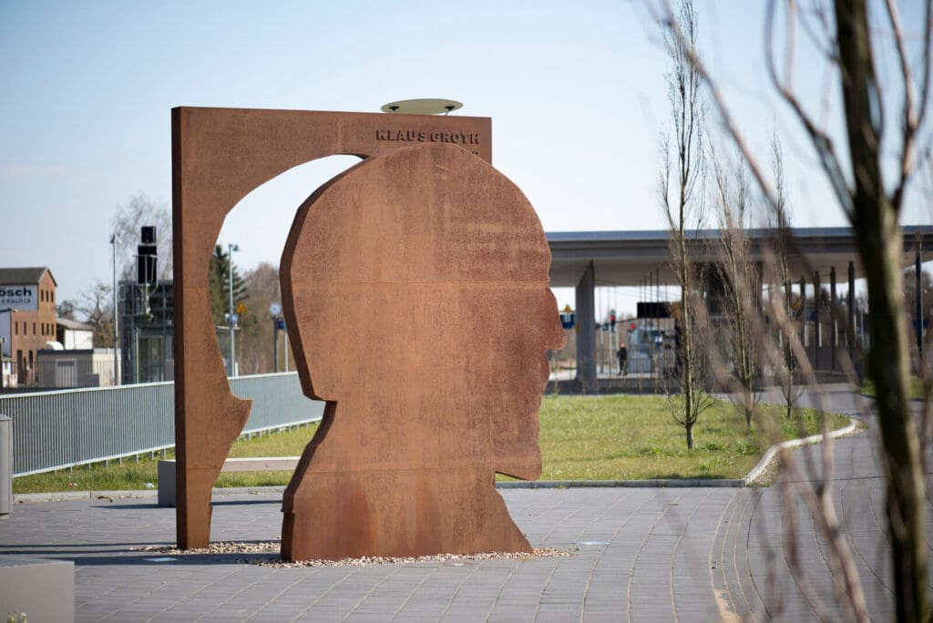 Auf dem Foto sieht man das großformatige Klaus-Groth-Denkmal aus Cortenstahl von Jörg Plickat am Bahnhofsvorplatz in Heide.