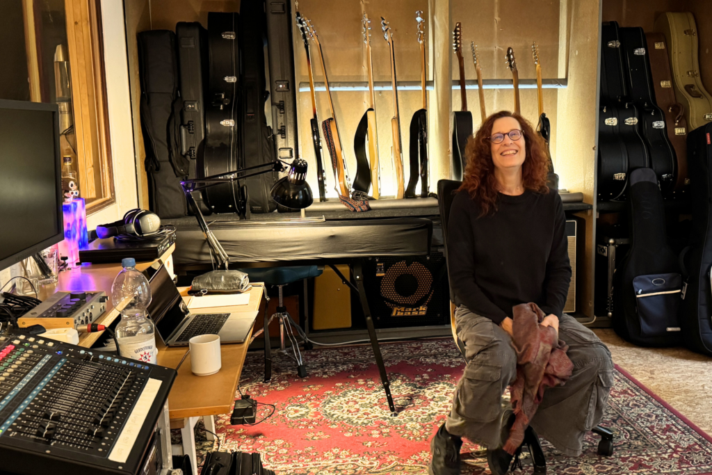 Auf dem Bild ist die Bassistin Susanne Vogel zu sehen. Sie sitzt in der Mitte ihres Proberaums, sie lacht. Hinter ihr sind zahlreiche Bassgitarren und Gitarren und die zugehörigen Koffer zu sehen, neben ihr Mischpult, Laptop und weiteres Equipment.